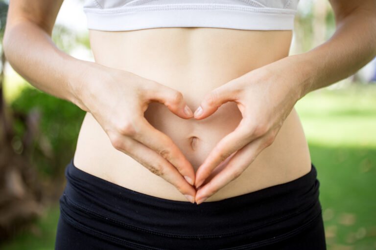 Female hands shaping heart on belly outdoors.