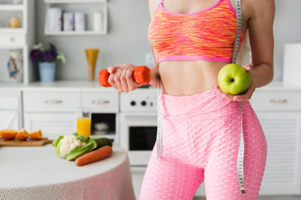 Fit woman in her kitchen holding an apply in one hand and dumbbell in other while showing off her skinny stomach