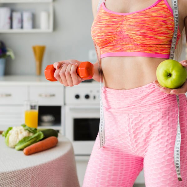 Fit woman in her kitchen holding an apply in one hand and dumbbell in other while showing off her skinny stomach