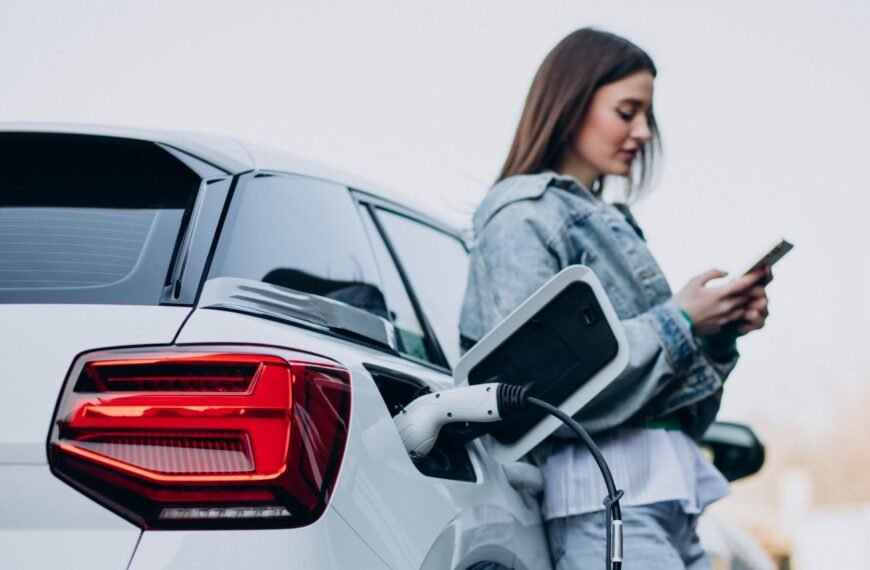 Young woman looking at her phone while charging her white electric car