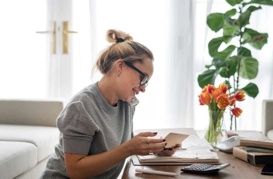 Woman in her living room at her desk talking on a phone and looking at paperwork