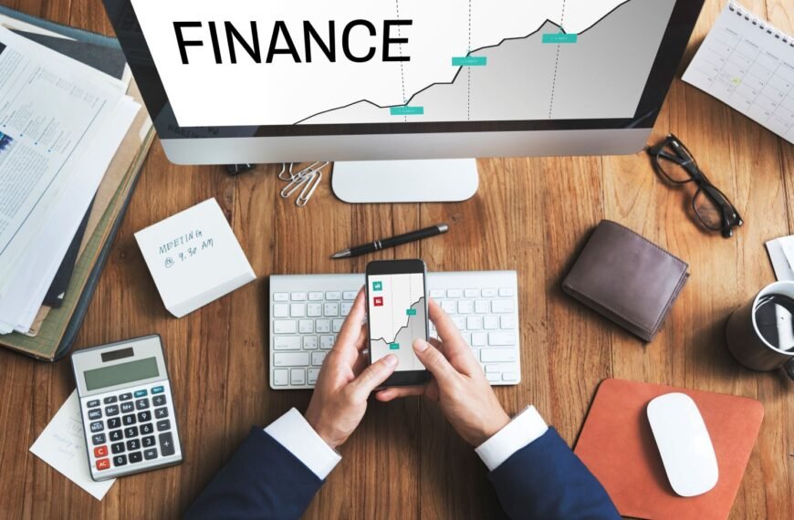 Man's hand over his desk holding a phone showing a graph with a computer screen in front of him that says finance.