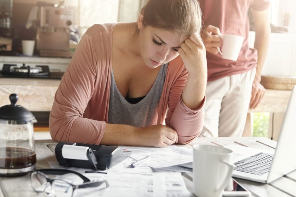 A woman in her kitchen table looks stressed about money while looking at papers and calculator in front of her. A man is standing behind her holding coffee cup